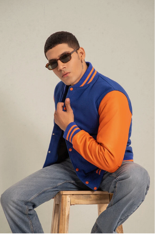 Man wearing a blue and orange varsity jacket sitting on a wooden stool.