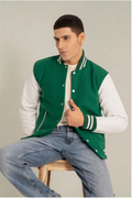 Man wearing a green and white varsity jacket sitting on a stool against a plain background
