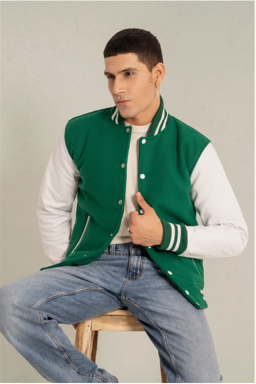 Man wearing a green and white varsity jacket sitting on a stool against a plain background