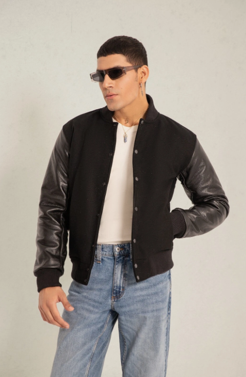 Man wearing a black varsity jacket with leather sleeves, white shirt, and blue jeans on a plain background