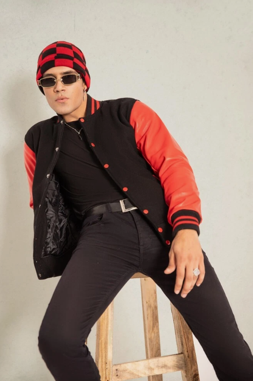 Person wearing a black and red varsity jacket, black pants, and a checkered hat sitting on a wooden stool.