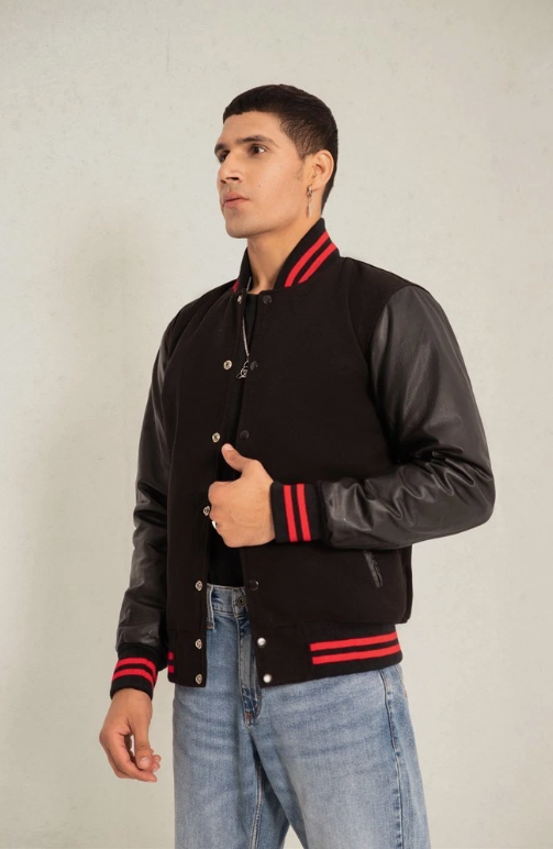 Man wearing a black varsity jacket with red accents and blue jeans on a plain background