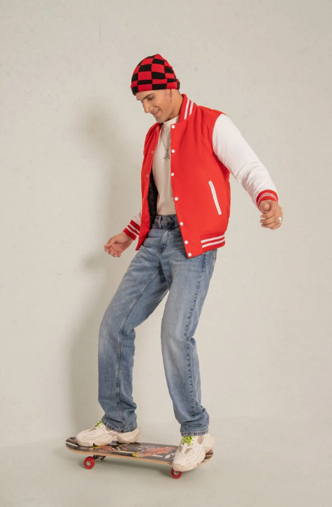 Man wearing a red varsity jacket, blue jeans, and a checkered beanie skateboarding on a plain background